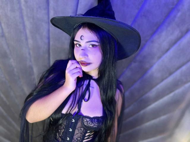 HannahBecker with black witch hat, long black hair, purple makeup, lace-up corset, and red lipstick, holding her lip, blue light, and gray fabric background. Boobs visible.