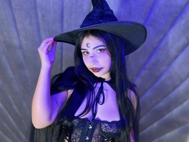 HannahBecker with black witch hat, long black hair, dark lipstick, white face paint, lace corset, holding hat, purple blue lighting, semi-sheer black top, exposing small tits.