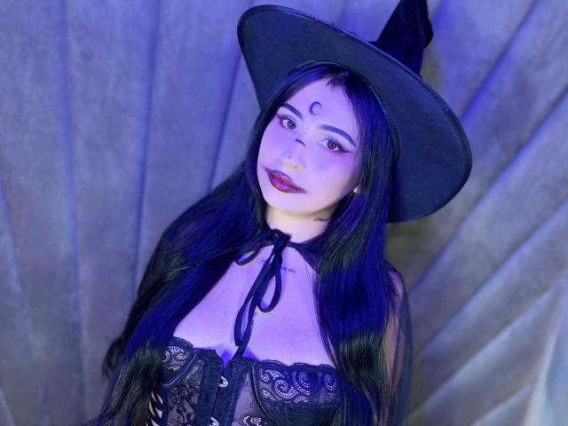 HannahBecker with long black hair, witch hat, black lace corset, blue eyes, red lipstick, moon symbol on forehead, tits visible, soft purple-blue lighting, looking up.