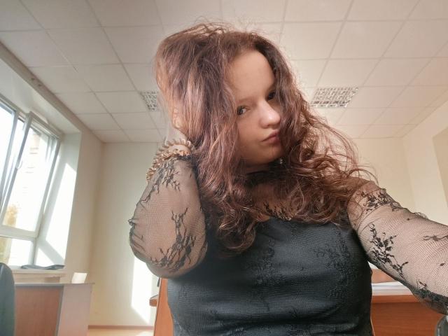 JinnyKosler with curly brown hair, wearing a black lace long-sleeve top, hands in hair, looking slightly to the side, in a bright room with windows.