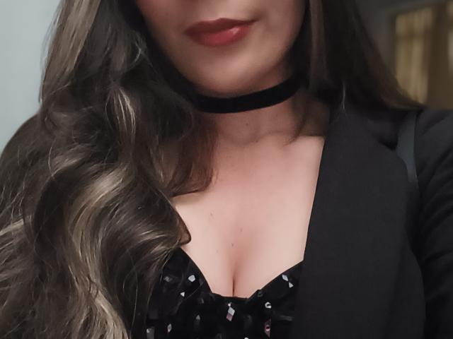 TaniaSandersX with wavy brown hair, red lipstick, black choker, black dress with pattern, showing cleavage, wearing black blazer, soft lighting, slightly smiling.