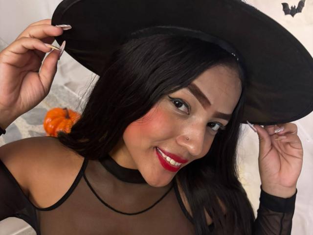 SheilaBrunette com um chapéu de bruxa preto, segurando a aba, sorrindo, batom vermelho, blusa preta transparente, colar, piercing no nariz, cabelo preto longo e uma abóbora laranja ao fundo. Ela é fofa, com seios..