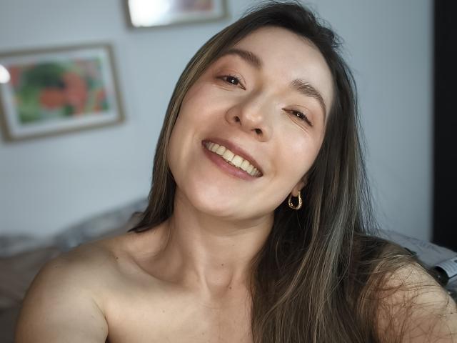 TaniaSandersX has long brown hair, smiling with white teeth, wearing gold earrings, and showing bare shoulders, looking happy in a selfie.
