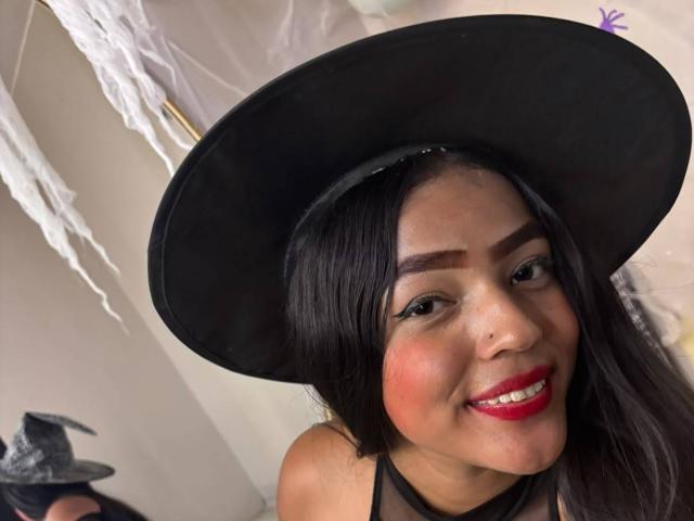 SheilaBrunette con cappello nero, rossetto rosso e eyeliner nero, sorridente, che mostra il piercing al naso, e indossa una maglietta nera, con il seno appena visibile..