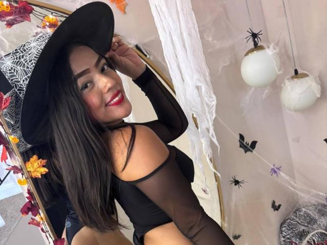 SheilaBrunette mit langen schwarzen Haaren, rotem Lippenstift, einem schwarzen, transparenten Oberteil und einem schwarzen Hut, lächelnd in die Kamera, wobei ihr Hintern zu sehen ist, trägt einen schwarzen Hexenhut, in einem Halloween-Themenzimmer..