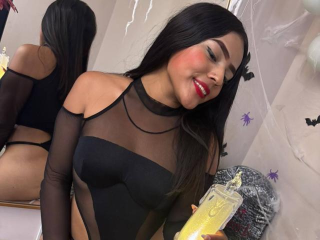 SheilaBrunette with long black hair, red lipstick, and green eyeshadow, wearing a sheer black top, smiling, holding a yellow drink, showing her ass in a mirror reflection.