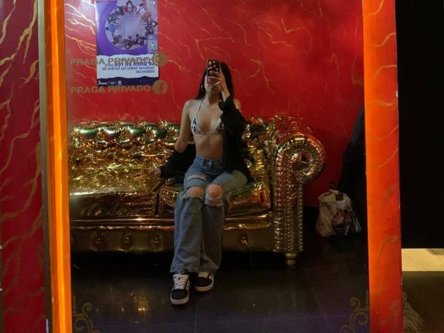 HannaThorne, mirror selfie, wearing black jacket, ripped jeans, blue bra showing small tits, black sneakers, taking picture with phone, sitting on gold couch, red wall background.