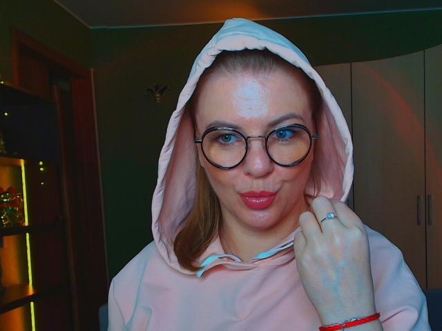 VeronikaGoldy with a shiny forehead, wearing a pink hoodie, glasses, and a ring, making a pouty face, blonde hair under the hood, blue eyes, in a dimly lit room.