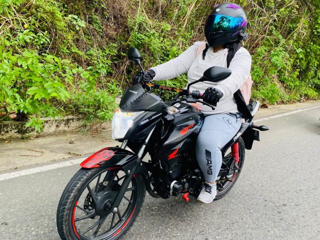 Melannie Carter fährt eine schwarze Motorrad mit roten Akzenten, trägt ein weißes Hemd, graue Hosen, schwarze Handschuhe und einen glänzenden, blauen, reflektierenden Helm auf einer asphaltierten Straße..
