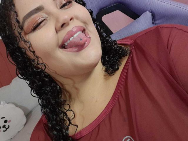 MelannieCarter with curly black hair, pink eyeshadow, and tongue out, showing a tongue piercing, wearing a red shirt, sitting in a purple chair.