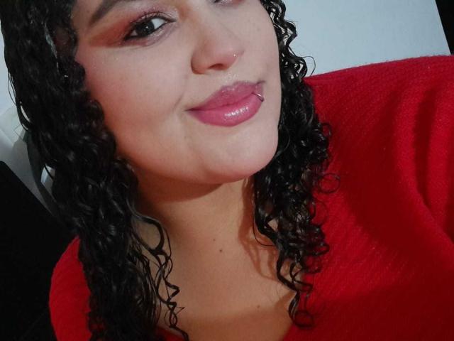 MelannieCarter with curly black hair, red lips, nose ring, and pink eye makeup, wearing a red top, smiling at the camera. Her tits are partially visible.