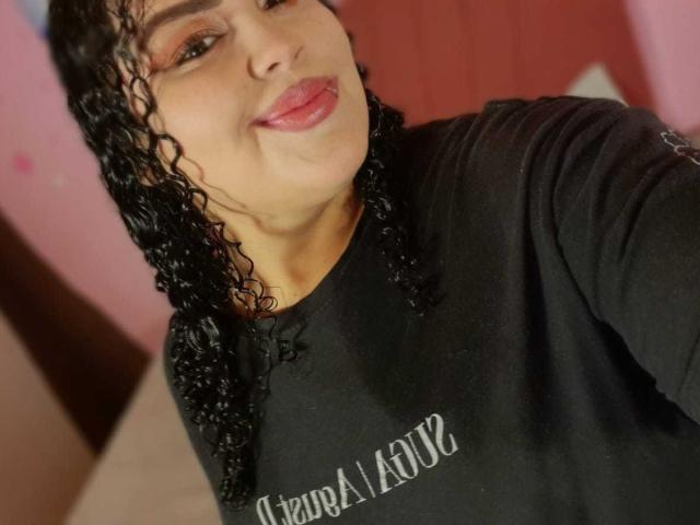 MelannieCarter with curly black hair, light brown skin, and pink lipstick, wearing a black shirt, smiling slightly, her small tits visible under the shirt.