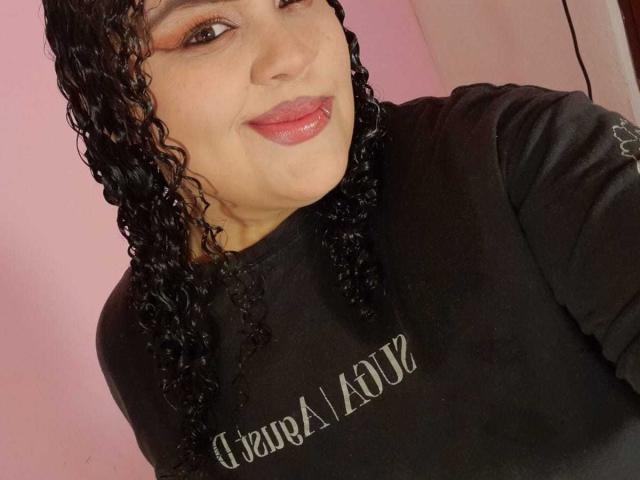 Melannie Carter mit lockigem, schwarzem Haar, pinkem Lippenstift und schwarzem T-Shirt, die ihre Zunge herausstreckt und im Hintergrund ihr leicht pralles Hinterteil zeigt. Weiches, rosa Licht..