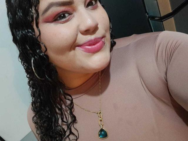 MelannieCarter has curly black hair, pink eyeliner, glossy pink lips, and a cute smile. She's wearing a pinkish-brown top and a gold necklace with a green teardrop pendant.