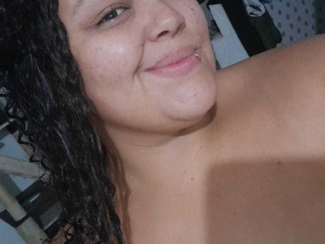 Melannie Carter mit lockigem, schwarzem Haar, gebräunter Haut und einem silbernen Piercing in der Lippe, lächelnd, wobei ihre nackten Schultern sichtbar sind, und mit kleinen Sommersprossen und einem leicht rundlichen Gesicht..