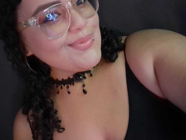 MelannieCarter with curly black hair, glasses, black choker, and black tank top, smiling at the camera, showing her tits and nipple. Dark background, soft lighting.