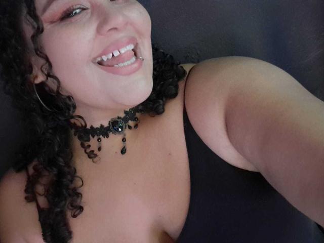 MelannieCarter with curly black hair, smiling with tongue out, black choker, black tank top, showing her tits, and a lip ring, against a dark background.