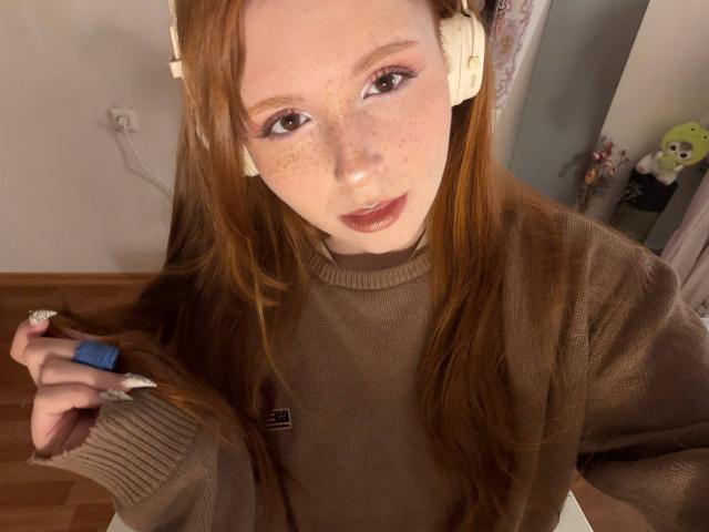 CorrinaGoodland with long red hair, freckles, and light makeup, wearing a brown sweater and white headphones, holding a lock of hair with a blue clip.