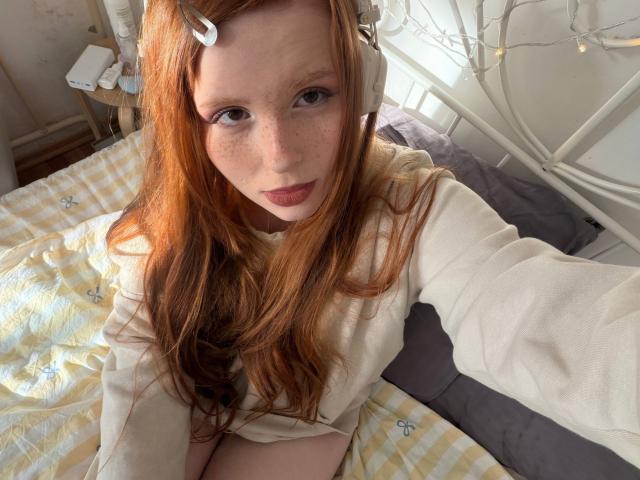 Corrina Goodland, avec ses cheveux roux, ses taches de rousseur et son casque blanc, portant un sweat crème, assise sur un lit jaune à carreaux, prend un selfie, montrant légèrement ses seins..