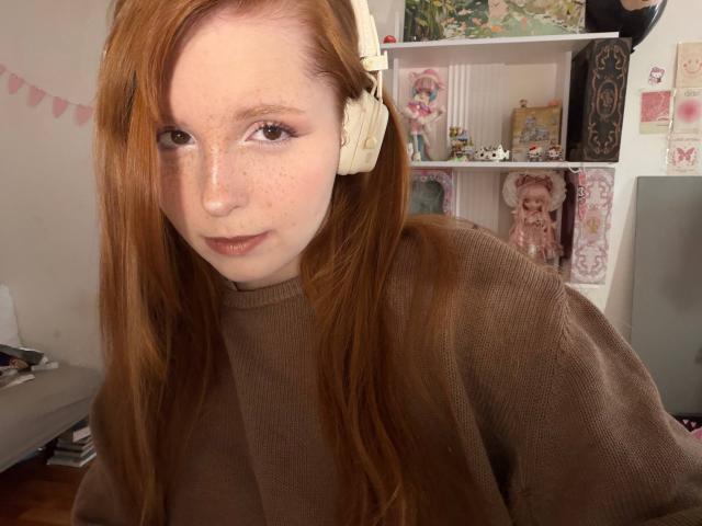 CorrinaGoodland with red hair, freckles, and brown eyes, wearing beige headphones and a brown sweater, looking at the camera with a subtle smile. Her tits are partially visible.