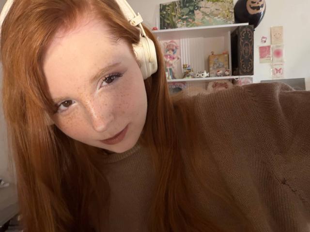 CorrinaGoodland has red hair, white headphones, freckles, brown sweater, and a slight smile. Her brown eyes look at the camera, showing a cute, relaxed expression.