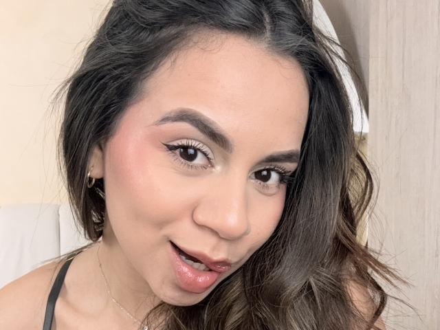 EmaRusso has wavy brown hair, thick eyebrows, pink blush, and is looking at the camera with a slight smile, wearing a black strap, and a thin necklace.