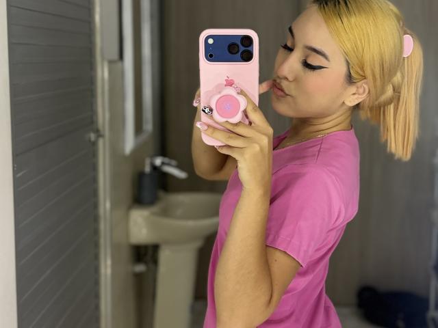EmmaWat with blonde pigtails, pink t-shirt, holding pink phone with heart grip, taking selfie in bathroom, pouty lips, eyeliner, pink nails, fair skin, cute, flirty expression.