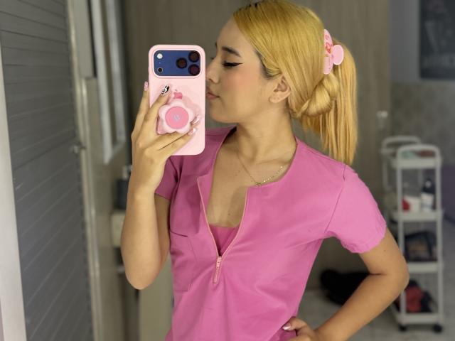 EmmaWat with blonde pigtails, pink scrubs, kissing phone camera, pink pop socket, small tits, pink nails, hand on hip, mirror selfie, pink phone case, in bathroom.
