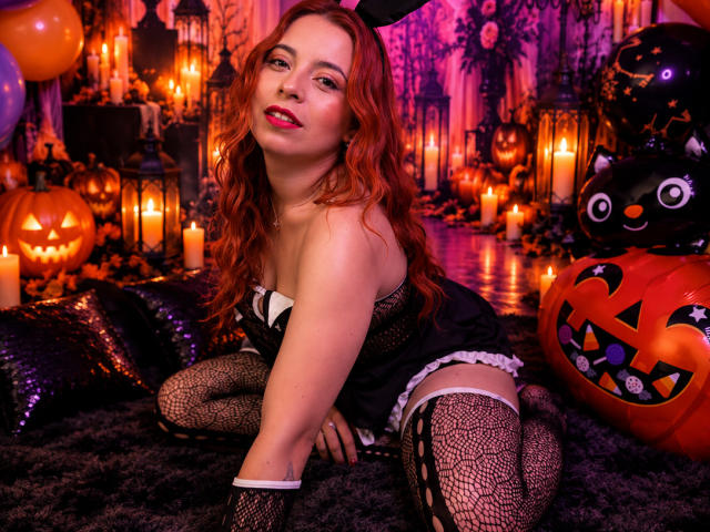MeghanStorm69, con cabello rojo, lencería negra y medias de red, arrodillada sobre una alfombra oscura, rodeada de velas encendidas y decoraciones de Halloween, mostrando sus senos y una sonrisa sensual..