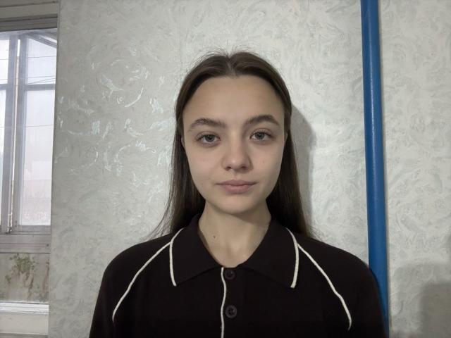 AbbeyBragan, com cabelo castanho liso, olhos castanhos e pele clara, vestindo uma camisa preta com detalhes brancos, está em frente a uma parede branca com textura..