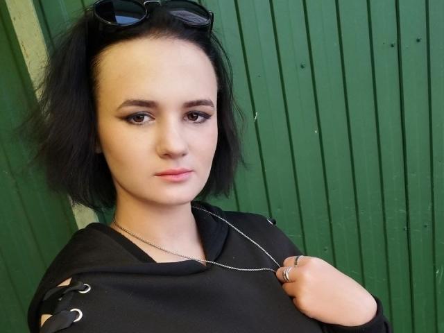 TreenaVisosky with short black hair, dark eyeliner, black top, silver necklace, sunglasses on head, green background, holding top with right hand, slight smirk, pale skin, cute.