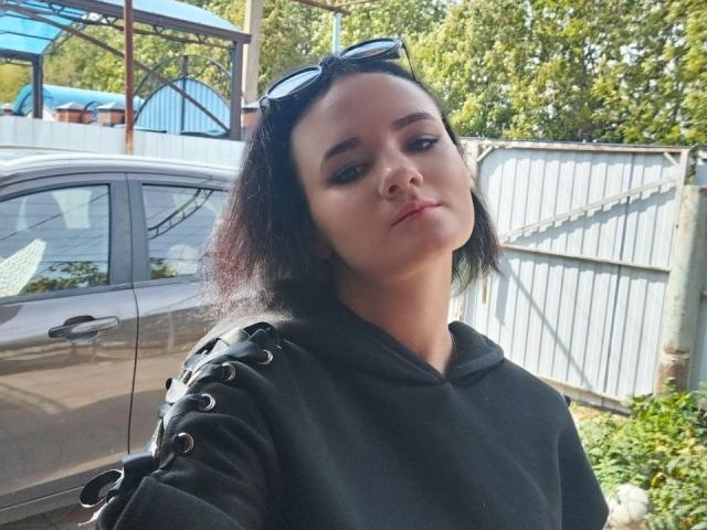 TreenaVisosky with short black hair, dark hoodie, and black straps on her shoulder, standing next to a brown car with sunglasses on her head, looking slightly to the side.