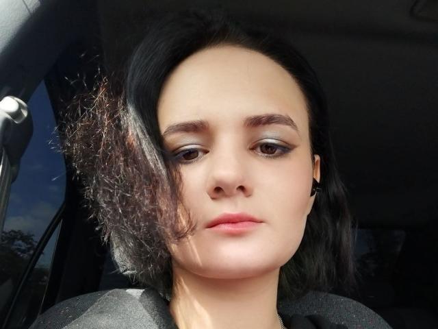 TreenaVisosky's face, with curly dark hair, light skin, and subtle makeup, looking straight ahead in a car, wearing a black shirt and small hoop earrings.