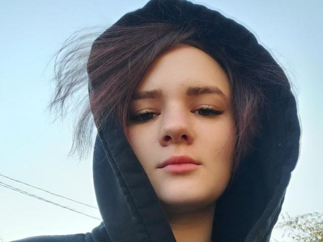 TreenaVisosky with messy dark brown hair, wearing a black hoodie, has light skin, half-closed eyes, and pink lips. She's looking down with a slightly bored expression.