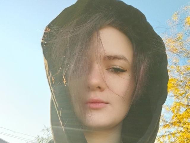 TreenaVisosky has pale skin, green eyes, and a serious expression, with messy brown hair covering one eye. She's wearing a black hooded jacket, and sunlight filters through.