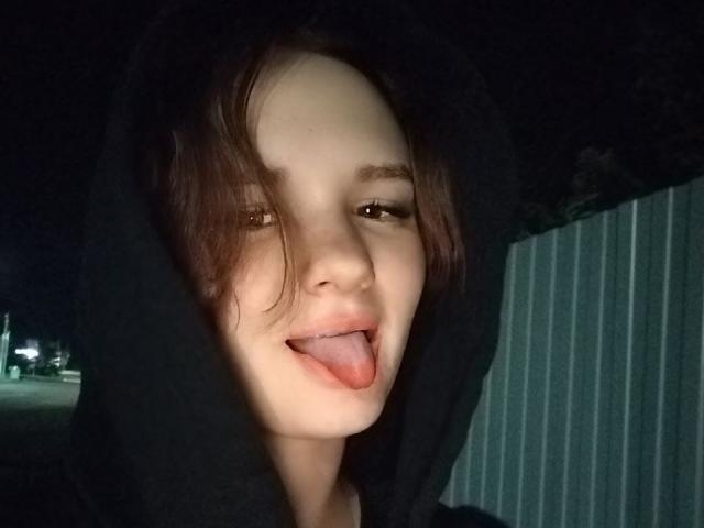 TreenaVisosky with pale skin, brown eyes, and messy brown hair, wearing a black hoodie, sticking out her tongue, in a nighttime selfie. Her tongue is slightly pink.