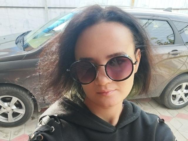 TreenaVisosky with curly dark hair, round sunglasses, black hoodie, and a serious face, standing in front of a brown car with silver wheels.