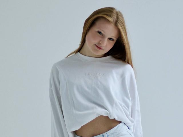 AkilahGhil with long blonde hair, wearing a white oversized shirt that exposes her midriff and part of her white panties, looking down with a slight smile.