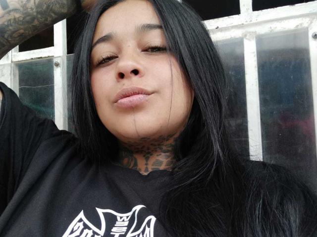 BlackRousse has long black hair, tattoos on her neck, and is wearing a black shirt with a white graphic. She's smirking with slightly parted lips.
