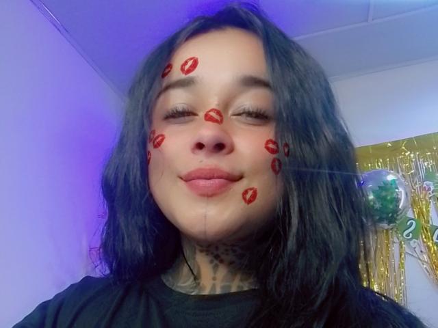 BlackRousse has red lipstick kiss marks on her face, smiling with eyes closed, black wavy hair, black shirt, and tattoo on her neck. Blue light in the background.