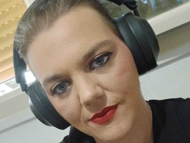 LaSoumise69, con gli occhi azzurri, il rossetto rosso, le cuffie nere e i capelli castani raccolti, sorride leggermente alla telecamera. Indossa una maglietta nera..