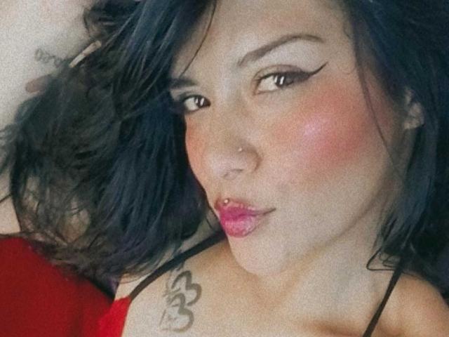 Mia Bellucci, con cabello azul oscuro, lápiz labial rosa, un piercing en la nariz y delineador de ojos negro, frunciendo sus labios rosados, mostrando un tatuaje en su hombro derecho, y vistiendo una blusa roja..