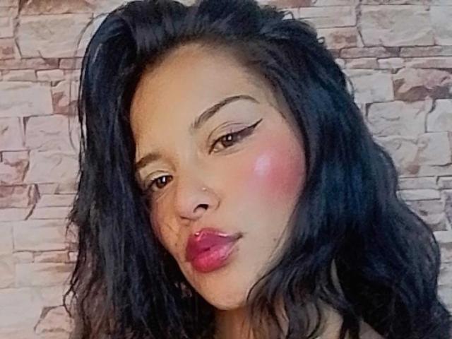 Mia Bellucci tiene el cabello oscuro y ondulado, lápiz labial rojo, delineador de ojos alado y un rubor rosado en las mejillas, mirando ligeramente hacia un lado con un leve puchero..