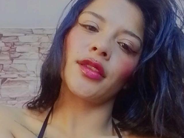 Mia Bellucci tiene el pelo azul, lápiz labial rosa y un pequeño piercing en la nariz. Está mirando ligeramente hacia un lado, con una expresión relajada y la boca ligeramente abierta. Sus senos no son visibles..