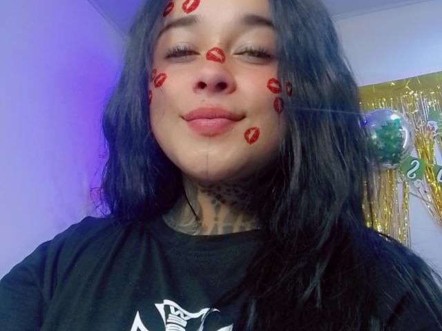 BlackRousse has red lipstick kiss marks on her face, with her eyes closed and a slight smile, wearing a black shirt with tattoos visible on her neck.