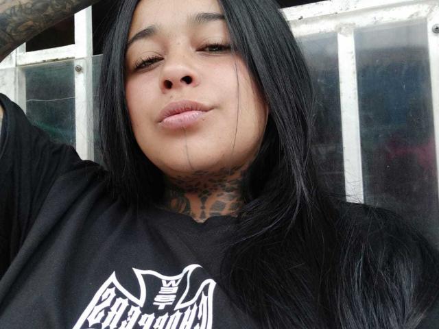 BlackRousse with long black hair, tattooed neck, and full lips, wearing a black shirt, winking slightly with a relaxed expression, and visible boob.