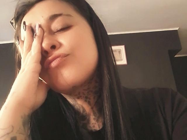 BlackRousse with dark hair, pensive expression, hand on face, tattoos on neck and hand, wearing black shirt, slightly puckered lips, soft lighting, close-up shot.