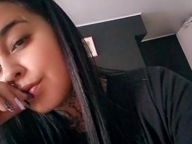 BlackRousse has long black hair, a tattoo on her neck, and is wearing a black shirt, with her finger near her slightly parted lips in a selfie.