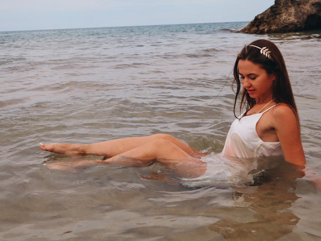KaraCriegs with wet white tank top, sitting in shallow water, legs up, brown hair, hair clip, looking down, arms behind her, visible pussy through wet top, rocky shore.