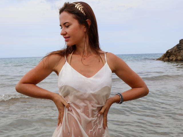 KaraCriegs in a wet, white, see-through dress, hands on hips, showing her small tits, with a bracelet on her right wrist, standing in shallow ocean water.